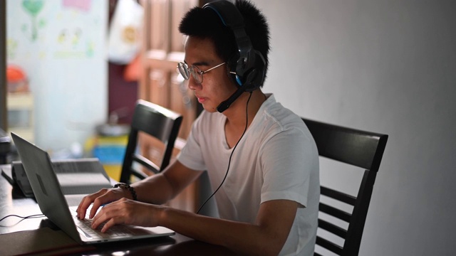 一名亚裔中国少年一边在厨房餐桌上做作业，一边通过网络软件与所有同学在线聊天视频素材