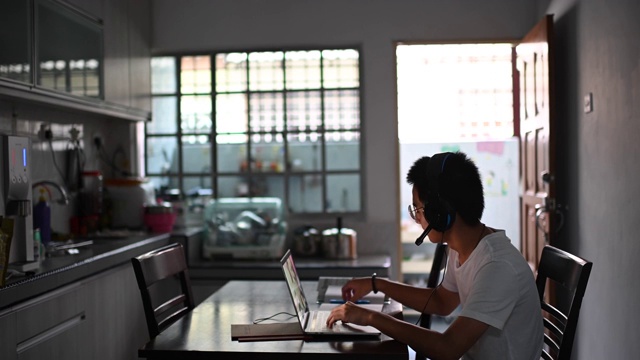 一名亚裔中国少年一边在厨房餐桌上做作业，一边通过网络软件与所有同学在线聊天视频素材