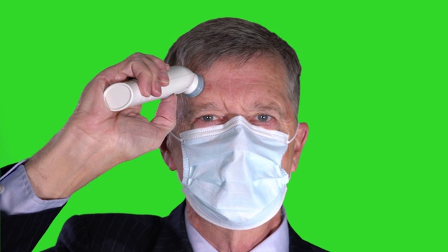 年长男子戴着口罩，在色度键绿色屏幕上测量自己的体温以检查病毒视频下载