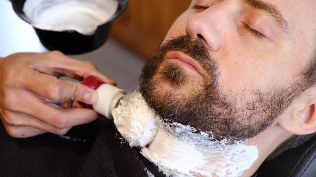 专业的美发师在理发店里为顾客涂抹剃须泡沫年轻人在理发店里把没刮胡子的胡须剪成形状。男性美容理念视频下载
