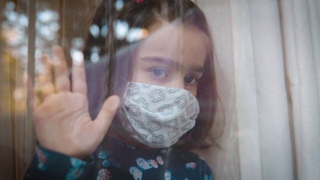 看起来很伤心的小女孩戴着防护面具透过窗户看，因为她不被允许出去玩。视频素材