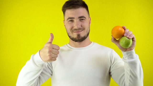 一个年轻人手里拿着一个新鲜的橘子和青苹果。一个穿着白色运动衫的男人露出一个超级标志和微笑。适当营养的概念。黄色背景简单视频素材