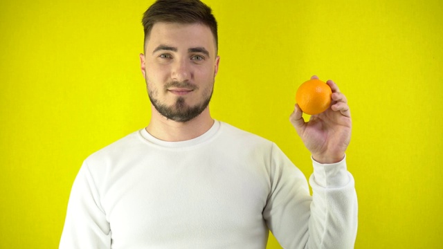 一个年轻人手里拿着一个新鲜的橘子。一个穿着白色运动衫的男人对着眼睛眨了眨眼睛，然后笑了。适当营养的概念。黄色背景简单视频素材