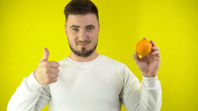 一个年轻人手里拿着一个新鲜的橘子。一个穿着白色运动衫的男人露出一个超级标志和微笑。适当营养的概念。黄色背景简单视频素材