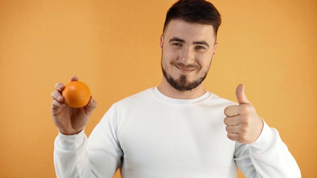 一个年轻人手里拿着一个桔子。一个穿着白色运动衫的男人微笑着，表现出一种超级的迹象。适当营养的概念。橙色简单的背景视频素材