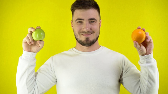 一个年轻人一手拿着桔子，另一手拿着青苹果。一个穿着白色运动衫的男人微笑着。适当营养的概念。黄色背景简单视频素材