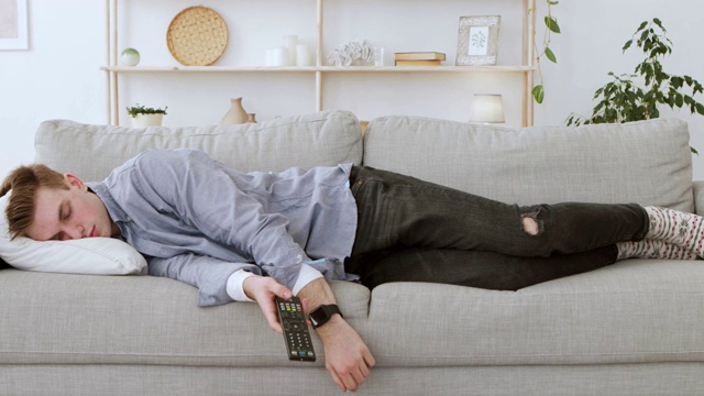 一个年轻人拿着遥控器在家里的沙发上打盹视频素材