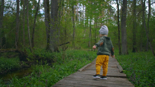 一个男孩在公园的一条木制小路上慢镜头奔跑。一个男孩在森林里的冒险。一个2-3岁的男孩在森林里跑过一座木桥。视频素材