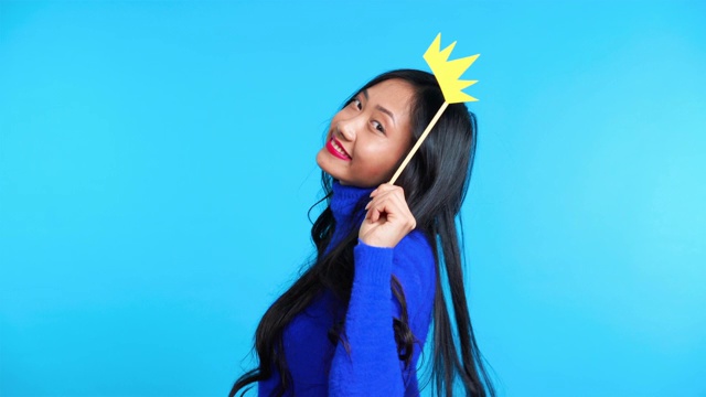美丽的亚洲女人与纸皇冠在棍摆姿势在蓝色的背景视频下载