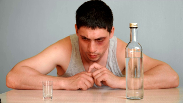 一个眼睛青肿的年轻人正在酗酒。一个男人喝醉了坐在家里的桌子上拿着一瓶酒精饮料。试图戒掉习惯。酗酒问题。撤军的痛苦。视频下载