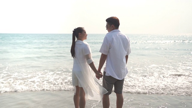 日落时分，一对年轻的亚洲男女手牵着手，微笑着一起走在沙滩上的慢镜头。幸福的家庭，丈夫和妻子放松，享受浪漫的暑假。视频素材