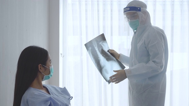 医生戴着面罩、医用口罩防护服、护目镜、呼吸面罩和手套，手持装有血样的试管，向生病的亚洲女性患者解释新冠肺炎疫情视频下载