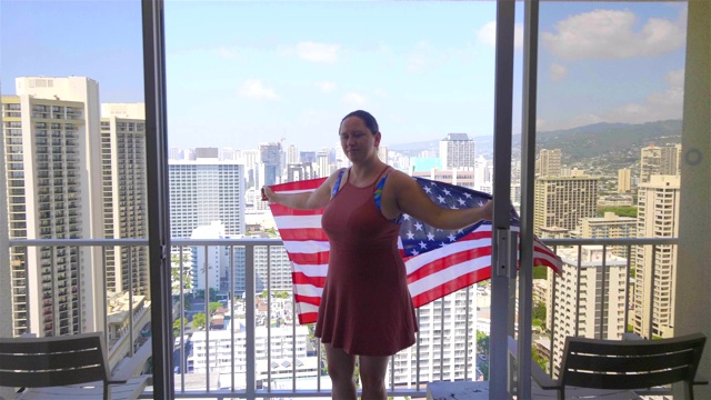 一个拿着美国国旗的女人站在阳台上，看着夏威夷檀香山的怀基基海滩，4k慢镜头60帧/秒视频下载