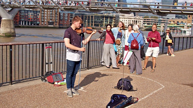 在河边为游客演奏小提琴的小提琴艺术家。远处的步行桥。伦敦金融区。视频下载