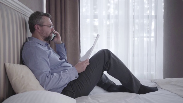资深商人坐在酒店房间的床上讲电话。自信的成年白人男子在旅行中戴着眼镜解释商业策略。旅行,生活方式,创业。视频下载
