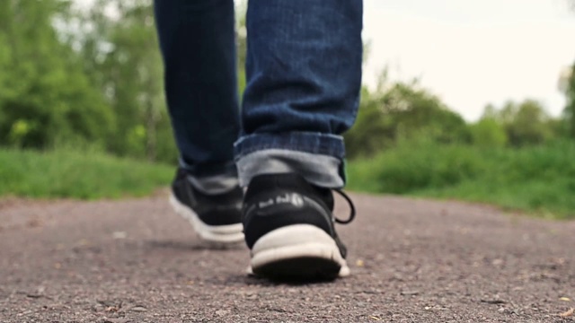 一个穿着运动鞋的男人走在一条土路上。穿着牛仔裤和运动鞋的男孩正在穿过公园。特写腿，一个人在走视频下载