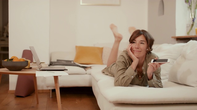 一个女人躺在沙发上用智能手机聊天，在家工作视频素材