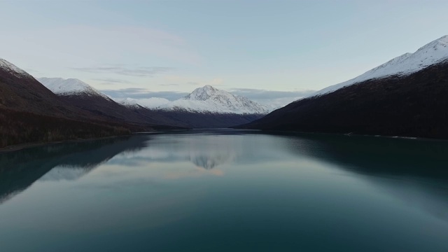 无人机摄像头盘旋在一个镜面湖上，湖面位于山顶积雪和山脚下的森林之间(美国阿拉斯加州埃克鲁特纳湖)视频素材
