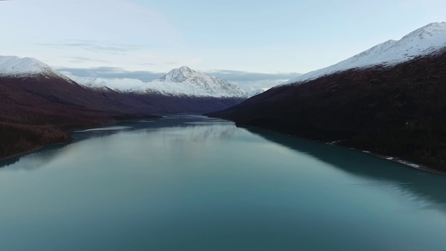 无人机平稳地离开一条平静的河流，在树木繁茂的山脉和积雪覆盖的山峰之间(埃克鲁特纳湖，阿拉斯加，美国)视频素材
