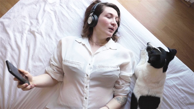 幸福的女人戴着耳机，躺在家里的床上用智能手机视频下载