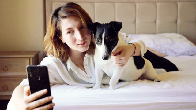 爱的女人亲吻可爱的小狗，在床上用智能手机自拍视频下载