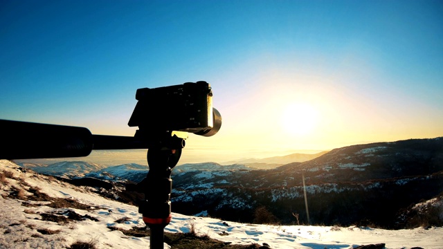 山顶三脚架上的剪影相机拍摄冬季日落的延时照片视频素材