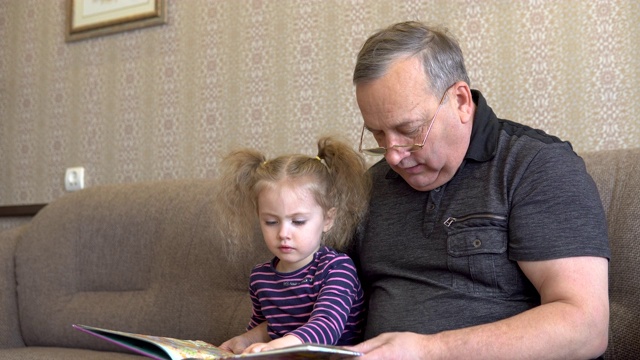 孙女正在和爷爷一起看书。女孩对着书皱着眉头，认真地听着爷爷的话。一起坐在沙发上特写视频素材