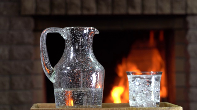 在燃烧的壁炉前的桌子上，有一个水罐和一杯带冰的矿泉水旋转着。视频下载