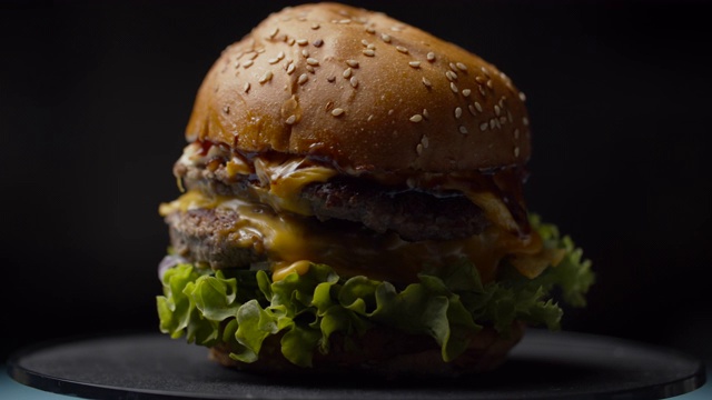 美味的汉堡与双层牛肉片，融化的奶酪，生菜和蔬菜旋转在黑暗的背景慢动作。视频素材