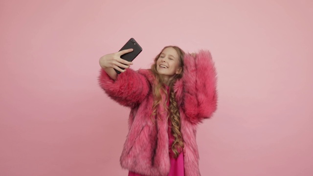一个年轻漂亮的女孩穿着粉红色的皮草大衣，在粉红色的背景下拍了很多自拍和飞吻。视频素材
