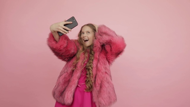 一个年轻漂亮的女孩穿着粉红色的外套，粉红色的背景做了很多自拍用手机。视频素材