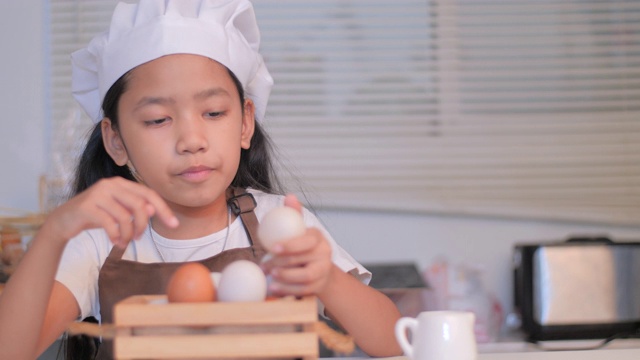 一个亚洲小女孩戴着一顶白色厨师帽，坐在厨房的木篮子里数鸡蛋视频素材