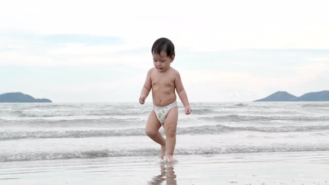 快乐的亚洲小男孩儿子，11个月大，在热带海滩上学习走路，沿着日落的边缘，在沙滩上冲浪。假期的概念。视频下载