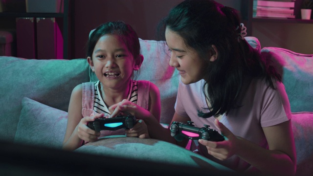 迷人的亚洲姐妹女孩在家里的客厅里一起玩电子游戏。由于冠状病毒封锁，两姐妹在家中使用无线控制器。室内霓虹灯装饰视频下载