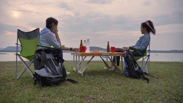 两个亚洲女子背包客在湖边欣赏夏日日落的慢镜头。他们是快乐的，有乐趣的假期，放松的时刻。青少年自然户外日常生活的概念视频素材