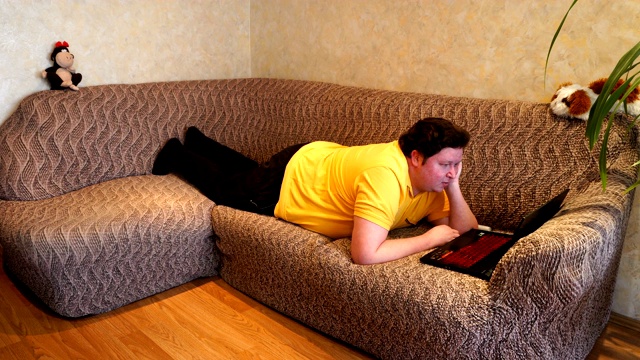 一个胖子躺在客厅的沙发上用笔记本电脑。年轻人上网，上网，看新闻视频下载