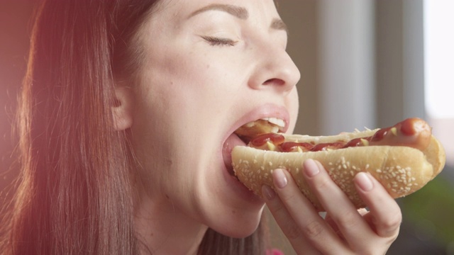 饥饿的女人吃热狗享受味道室内阳光照耀红色相机的特写镜头视频下载