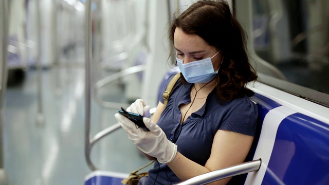 一名女性戴着医用口罩和防护手套，在地铁里旅行时沉迷于智能手机视频下载