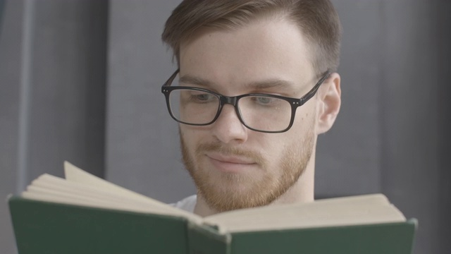 特写的聪明的年轻人在眼镜阅读的书。蓄着胡须的白人读者在室内欣赏文学作品。生活方式，爱好，智力，教育。电影4k ProRes总部。视频素材