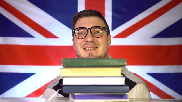 可爱的男学生戴着眼镜从一堆书中探出脸来，在英国国旗的背景下微笑着。英国的教育。教育的概念视频下载