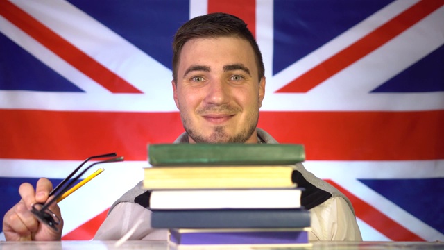 戴眼镜的可爱男生从一堆书中微笑着向外看，在英国国旗的背景下，他摘下了眼镜。英国的教育。教育的概念视频下载