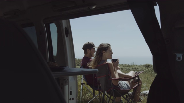 年轻的成年夫妇在公路旅行聊天和喝咖啡外露营车和看海景视频素材