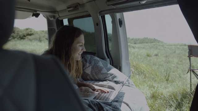 年轻的成年女性在露营车里用笔记本电脑在床上工作，看外面的风景视频素材