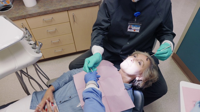 一名戴着N95口罩的男性牙医在一名60多岁的女性患者上方调整牙科灯，该女性患者躺在牙科诊所检查室的牙医椅上，牙科保健师在协助她摘下口罩视频素材