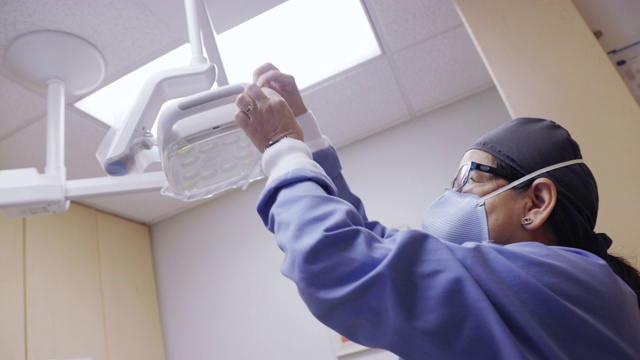 在牙科诊所的检查室里，一名50多岁戴口罩的拉丁裔保健工作者在牙科灯的把手上涂上干净的塑料罩视频素材