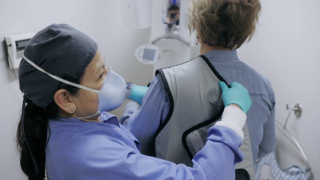一名50多岁的拉丁裔牙科卫生员戴着面罩，将保护铅围裙系在一名准备在牙科诊所拍x光片的妇女身上视频下载