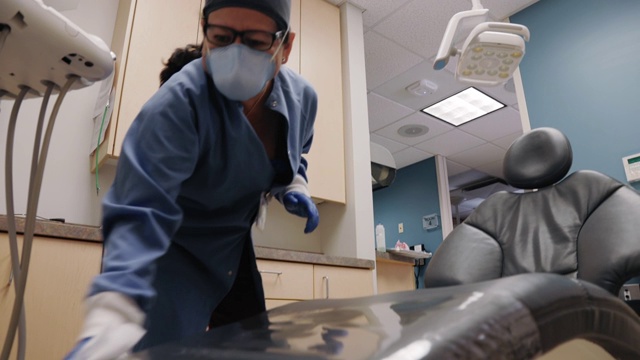 在牙科诊所的检查室里，一名50多岁的拉丁裔保健工作者戴着口罩和手术手套，在纸巾上喷洒消毒剂，擦拭牙医的椅子，为下一个病人做准备视频素材