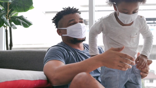 一个非洲家庭，父亲带着小男孩戴着医用口罩，待在家里预防新冠病毒或Covid-19的流行。父亲和小男孩在一起。视频素材