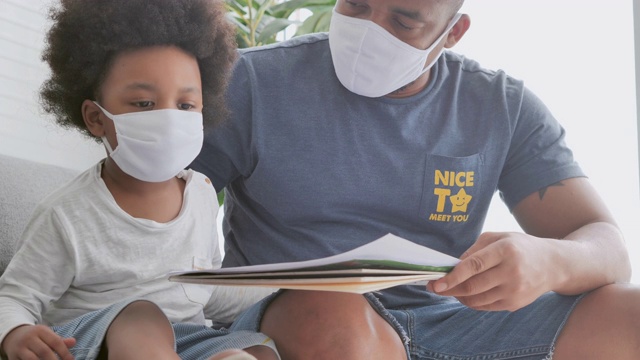 这是一个非洲家庭，父亲的小男孩在家上学时戴着医用口罩，为了预防新冠病毒或Covid-19的流行，他们呆在家里。父亲和小男孩在一起。口罩及面罩(非白种人)视频素材