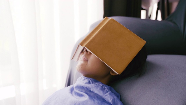 年轻的亚洲女人放松和睡着的时候，在客厅的沙发上看书，特写镜头。视频素材